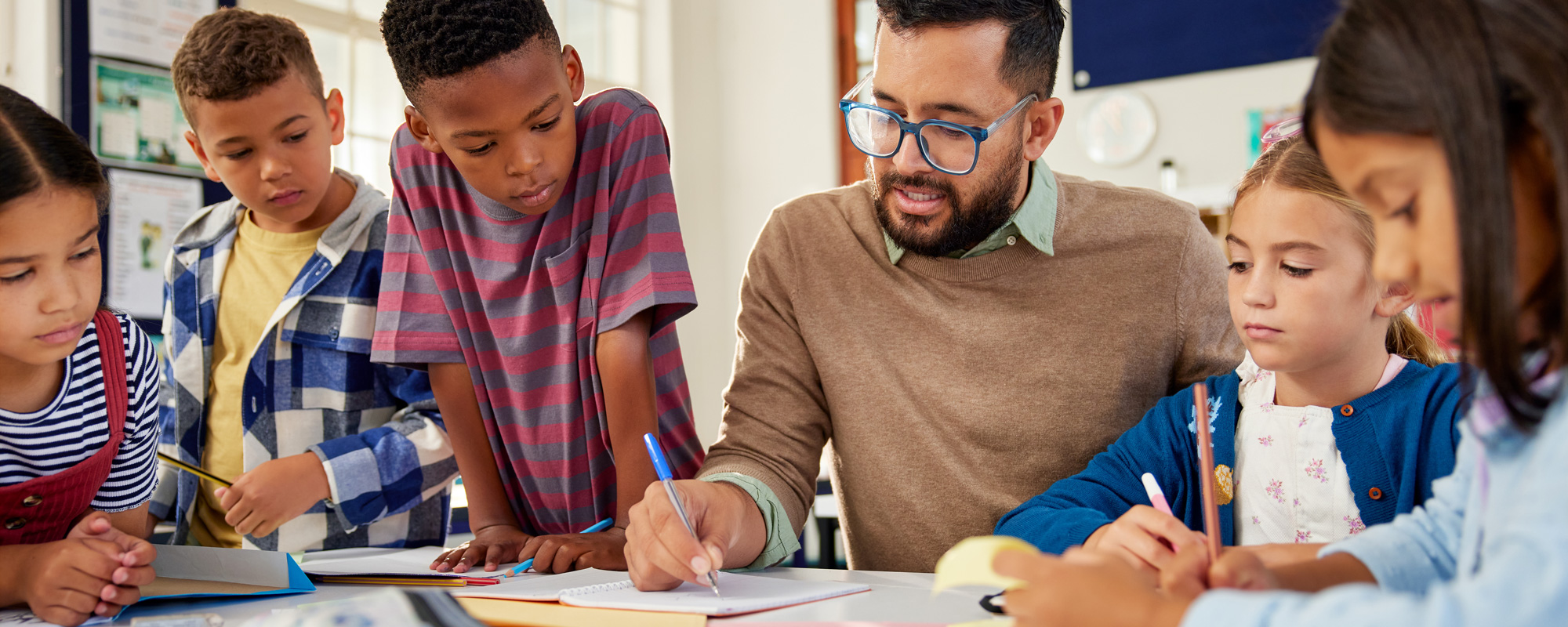 Male teacher sitting down working at a table with several young students