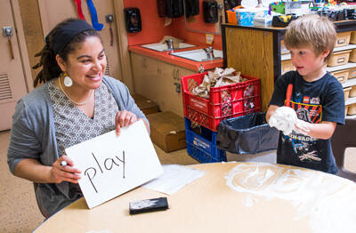 Teacher sitting at a table with a young student holding up a sign readying play